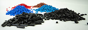 sechs verschiedenfarbene Häufchen Kunststoff-Granulate in den Farben :schwarz, blau, türkis, rot und braun liegen auf einem hellen Tisch 
